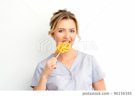 Portrait of a beautiful young caucasian beautician wearing a medical shirt bites a lollipop on a white background Portrait of a beautiful young caucasian beautician wearing a medical shirt bites a lollipop on a white background 90156385