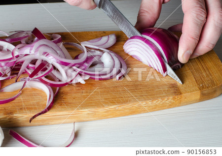 A man cuts red onions on a wooden chopping Board with a kitchen knife. Hands close up. Preparing food. Side view A man cuts red onions on a wooden chopping Board with a kitchen knife. Hands close up. Preparing food. Side view 90156853