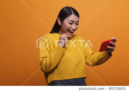 Young adult asian woman celebrating game victory on smartphone device. Happy playful positive person wearing yellow sweater celebrating winning mobile phone game while on orange background. Young adult asian woman celebrating game victory on smartphone device. Happy playful positive person wearing yellow sweater celebrating winning mobile phone game while on orange background. 90158693