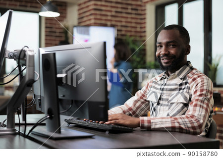 Agency adult happy smiling worker at computer in startup studio space modern interior. African american creative company employee sitting at desk in office workspace while looking at camera. Agency adult happy smiling worker at computer in startup studio space modern interior. African american creative company employee sitting at desk in office workspace while looking at camera. 90158780