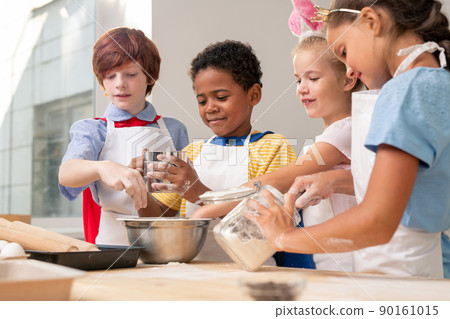 Multi-ethnic kids wearing aprons looking at each other while discussing festive menu while baking cookies 90161015