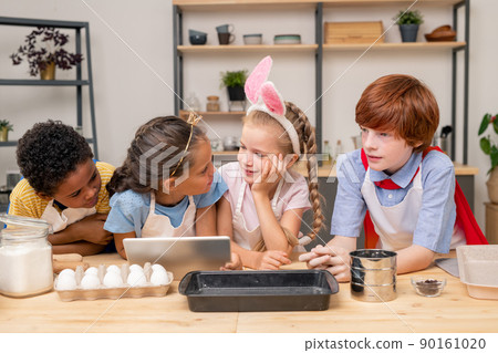kids wrapped up in reading recipe with help of digital tablet while standing at wooden kitchen table in order to make surprise for Mothers Day kids wrapped up in reading recipe with help of digital tablet while standing at wooden kitchen table in order to make surprise for Mothers Day 90161020