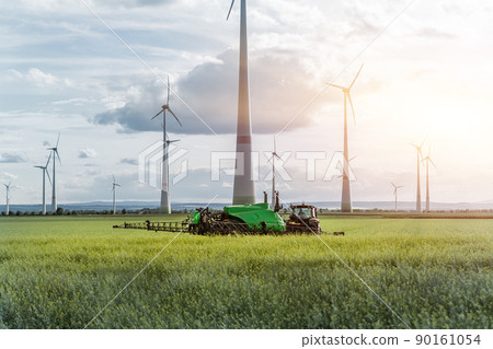 Scenic landscape view big modern tractor machine with sprayer equipment spraying fertilizer in rapeseed agricultural field against windfarm power windmill. Arable land cultivation sustainable energy 90161054