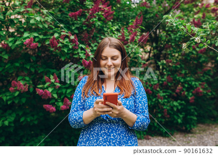 A portrait of a smiling beautiful person texting sms with her phone on nature background. Happy woman is using a smartphone in park outdoors, communications and technology concept 90161632
