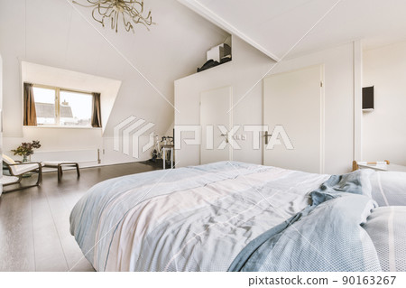 Comfortable bed located against window near wooden armchair and plant and doors in contemporary mansard bedroom 90163267
