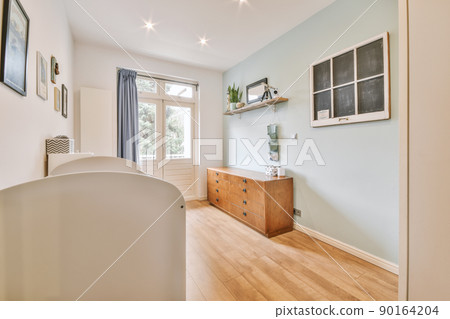 Interior of white room with baby crib and armchair near window with blue chest of drawers in daylight Interior of white room with baby crib and armchair near window with blue chest of drawers in daylight 90164204