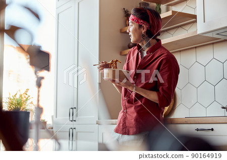 Smiling Asian guy leaning on the kitchen countertop and eating at home 90164919