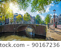 Famous romantic Amsterdam - beautiful sunny day on the old street of Amsterdam. Narrow canal, old bridge and traditional houses. Sunlight. Amsterdam, Holland, Netherlands, Europe. 90167236