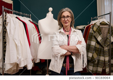 Confident elderly European woman fashion designer poses with crossed arms, looks at camera, standing near mannequin and new collection of women clothes on hangers 90169064