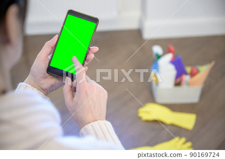 a woman holds a phone against the background of a plastic basket with detergents, brushes, sponges and rubber gloves for cleaning. a woman holds a phone against the background of a plastic basket with detergents, brushes, sponges and rubber gloves for cleaning. 90169724