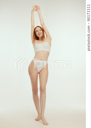 Full-length portrait of young slim red-haired girl posing in white underwear isolated over grey studio background 90171511
