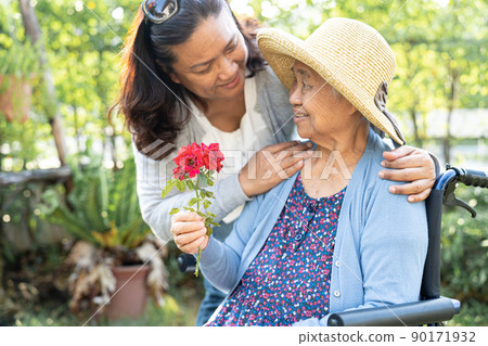 Caregiver daughter hug and help  Asian senior or elderly old lady woman holding red rose on wheelchair in park. 90171932
