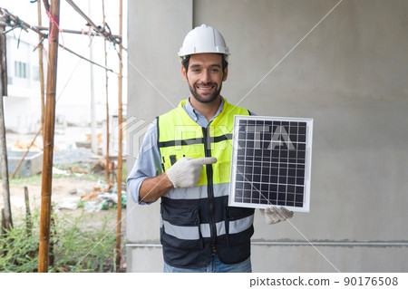 Young engineer in hardhat safety vest and glove pointing finger at solar cell panel, present a source of energy to generate direct current electricity with a smile. Young engineer in hardhat safety vest and glove pointing finger at solar cell panel, present a source of energy to generate direct current electricity with a smile. 90176508