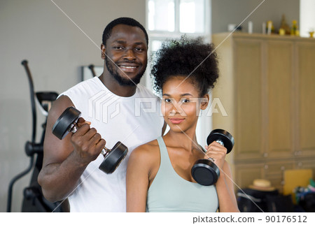 Young curly black hair couple enjoy holiday together in fitness center. Both lifting barbell with one hand. Young curly black hair couple enjoy holiday together in fitness center. Both lifting barbell with one hand. 90176512