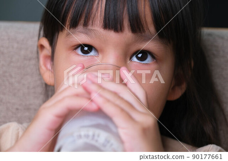 Portrait of a cute Asian little girl holding a glass of milk sitting on the sofa at home. Small girl at home with smiling face, feeling happy enjoying drinking milk and looking at camera. Portrait of a cute Asian little girl holding a glass of milk sitting on the sofa at home. Small girl at home with smiling face, feeling happy enjoying drinking milk and looking at camera. 90177661