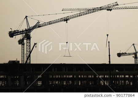A large construction site with busy cranes. Tower cranes working on a construction site lifts a load at high-rise building in evening. 90177864