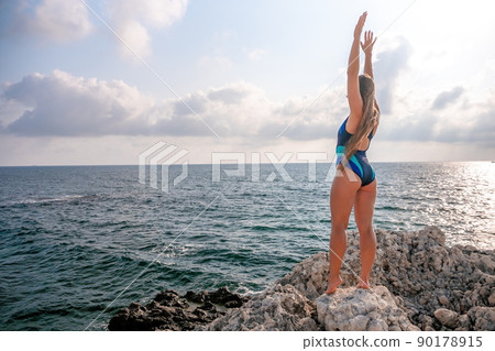 A young beautiful tanned woman with long hair in blue bikini stands at rock near blue green clear ocean sea water. Woman relax on sunset at sea, enjoy holidays and weekend vacation in summer time. A young beautiful tanned woman with long hair in blue bikini stands at rock near blue green clear ocean sea water. Woman relax on sunset at sea, enjoy holidays and weekend vacation in summer time. 90178915