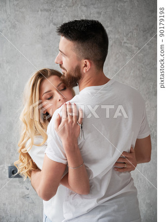 Woman holding house keys and embracing husband in new apartment. Happy couple hugging and celebrating real estate purchase. Concept of love, new home and property purchase. 90179198