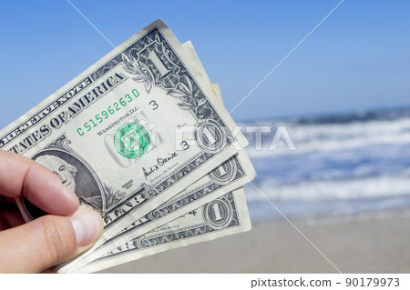 Person holding three paper dollar bills on background of sea and sea waves with white foam, sandy beach and clear blue sky on sunny summer day. Concept of money, expenses on vacation, travel, tourism 90179973