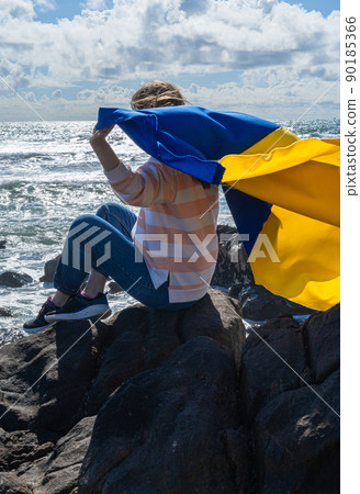 Refugee woman holding national flag of Ukraine sitting by the ocean. 90185366