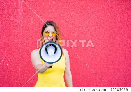 Indignant woman in sunglasses shouts into a megaphone on a red background. Indignant woman in sunglasses shouts into a megaphone on a red background. 90185689