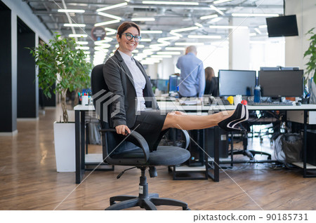 Happy business woman doing abdominal muscle exercises in an open space office. A red-haired smiling female employee in a skirt and high heels is dying off a chair in the workplace. Fitness at work. 90185731