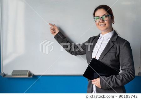 Beautiful woman in a suit with a digital tablet in her hands. A smiling female business coach with glasses stands in a conference room and points a finger at a white board. Workshop at the office. Beautiful woman in a suit with a digital tablet in her hands. A smiling female business coach with glasses stands in a conference room and points a finger at a white board. Workshop at the office. 90185742