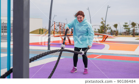 Caucasian woman in a mint sweatshirt is training with battle ropes at the sports ground.  90186484