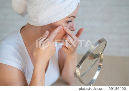 A Caucasian woman with a white terry towel on her head looks in the mirror and crushes pimple. Problem skin during menstruation. Acne and hyperandrogenism due to hormonal disorders. 90186490