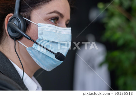Close-up of a woman in a medical mask talking on a headset in an office. Portrait of a female call center operator during a virus outbreak. Business worker answer customer calls during outbreak covid. Close-up of a woman in a medical mask talking on a headset in an office. Portrait of a female call center operator during a virus outbreak. Business worker answer customer calls during outbreak covid. 90186518