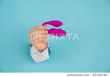 A woman's hand holds a pink sex toy through a hole in a paper blue background. A woman's hand holds a pink sex toy through a hole in a paper blue background. 90186790