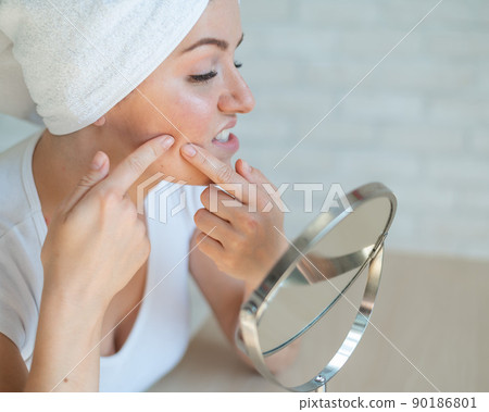 A woman with a white towel on head looks in the mirror and squeezes a pimple on cheek. A girl with hormonal problems does facial cleansing at home. Teen acne. Dermatologist himself in isolation. 90186801