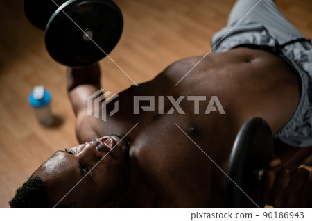 Shirtless afro american man doing exercises with dumbbells lying on bench in gym. Shirtless afro american man doing exercises with dumbbells lying on bench in gym. 90186943