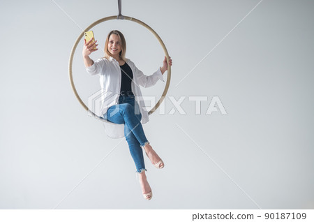 Caucasian woman gymnast on an aerial hoop takes a selfie on a smartphone. Caucasian woman gymnast on an aerial hoop takes a selfie on a smartphone. 90187109