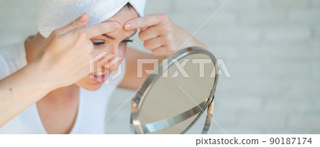 An upset woman looks in the mirror and squeezes a pimple on her forehead. A girl with hormonal problems cleanses her face after a shower. Teen acne. Himself a dermatologist. Problem skin. 90187174
