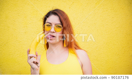 A beautiful red-haired woman in a yellow dress and sunglasses shows her tongue and and licks a banana. Portrait of a fantasizing girl with ripe fruit. A beautiful red-haired woman in a yellow dress and sunglasses shows her tongue and and licks a banana. Portrait of a fantasizing girl with ripe fruit. 90187176
