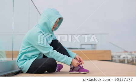 Caucasian woman in a hoodie tying her shoelaces before jogging. Caucasian woman in a hoodie tying her shoelaces before jogging. 90187177