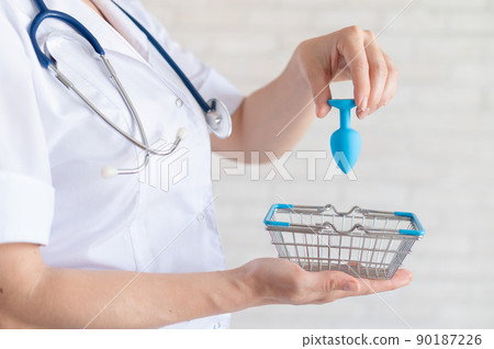 Faceless gynecologist puts anal plug in miniature shopping baskets. The doctor holds the device for alternative sex. A practitioner recommends a silicone toy for safety. 90187226