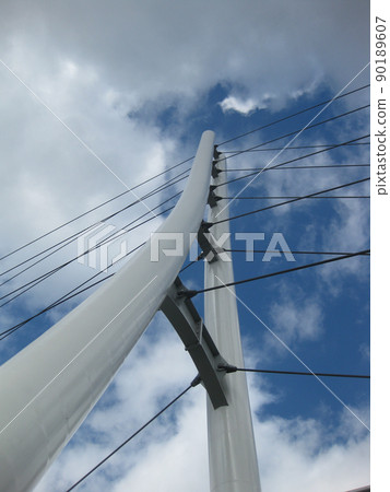Worm's eye view of a white triangular bridge with suspension cables crisscrossing a blue and cloudy sky Worm's eye view of a white triangular bridge with suspension cables crisscrossing a blue and cloudy sky 90189607