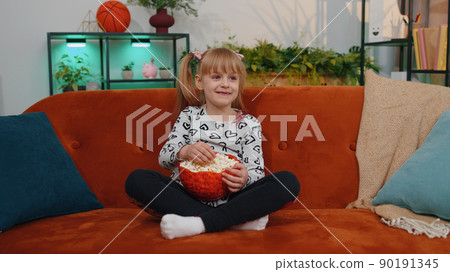 Funny children girl watching comedy video film on tv, eating popcorn on comfortable sofa at home Funny children girl watching comedy video film on tv, eating popcorn on comfortable sofa at home 90191345