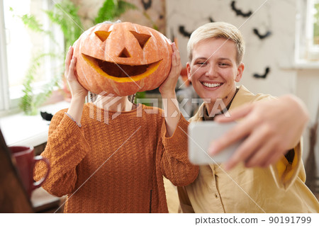 Smiling young woman with blond hair taking funny selfie with friend covering face with carved pumpkin at Halloween party 90191799