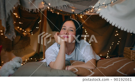 A young woman looks at the camera and laughs while lying in a tent of blankets and pillows in her bedroom. Leisure 90192081