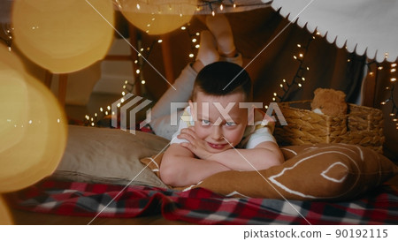 A little boy looks at the camera while lying in a tent of blankets and pillows in the bedroom. Children 90192115