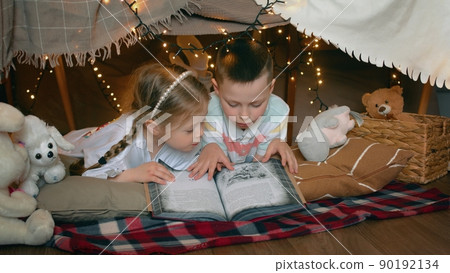 A little boy and girl are lying and reading a book in a tent of blankets and pillows in the bedroom. Children A little boy and girl are lying and reading a book in a tent of blankets and pillows in the bedroom. Children 90192134