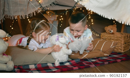 Little boy and girl play with toys and laugh while lying in a tent of blankets and pillows in the bedroom. Children Little boy and girl play with toys and laugh while lying in a tent of blankets and pillows in the bedroom. Children 90192137