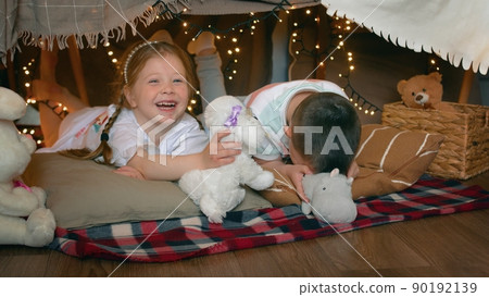 Little boy and girl play with toys and laugh while lying in a tent of blankets and pillows in the bedroom. Children Little boy and girl play with toys and laugh while lying in a tent of blankets and pillows in the bedroom. Children 90192139