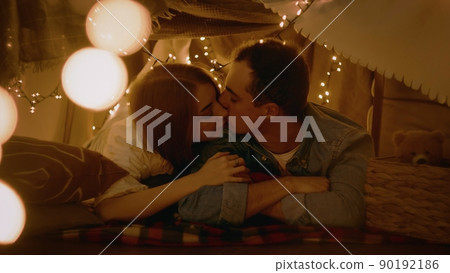 A young couple lies and kisses in a tent of blankets and pillows in the bedroom. Romance 90192186