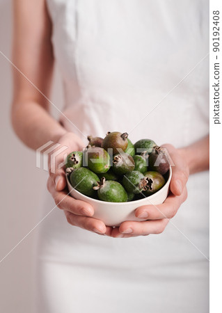 Vertical medium section shot of unrecognizable young woman wearing white dress holding bowl full of pineapple guava fruits, copy space 90192408