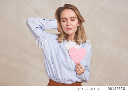 Horizontal medium portrait of young blond woman wearing casual outfit standing with eyes closed holding paper pink heart 90192858