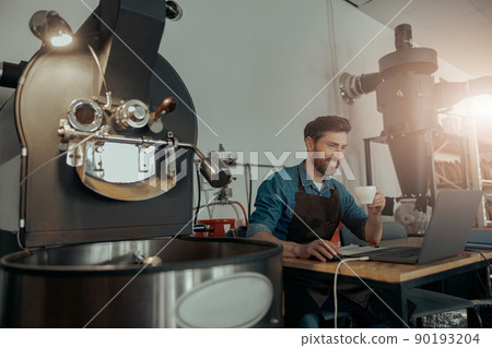 Barista on coffee roasting factory working on laptop and drinking coffee Barista on coffee roasting factory working on laptop and drinking coffee 90193204